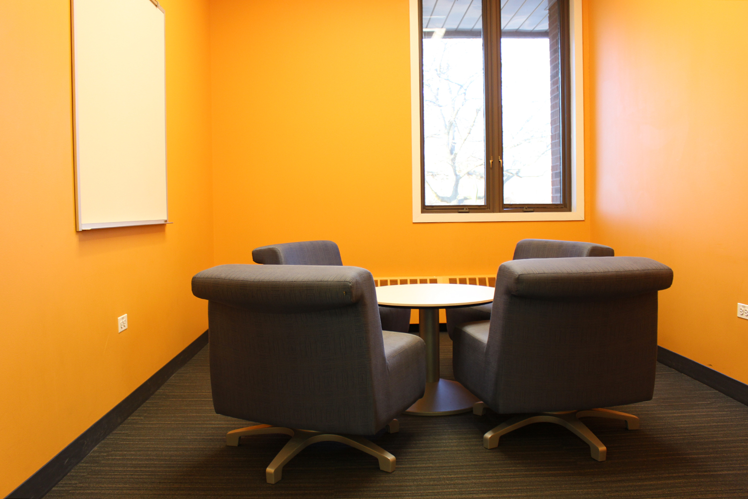 Room with orange walls, low table surrounded by four chair, white board on wall, window facing library parking lot