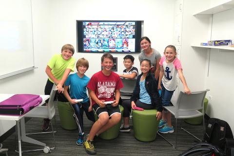 Youth Services Game Room interior shot with mounted television, various chairs, whiteboard, and a group of smiling children playing Super Smash Brothers Ultimate