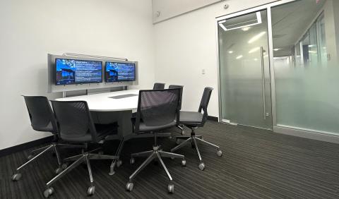 Small group meeting room with large glass doors, half circle table, gray chairs, two video display screens above table